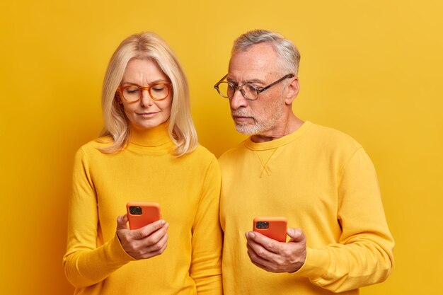 Zeitgenössisches älteres Ehepaar, das zuversichtlich ist, dass Technologiebenutzer sich aufmerksam auf Smartphones konzentrieren, überprüfen Informationen, tragen Brillen zur Sehkorrektur, die über der gelben Studiowand isoliert sind