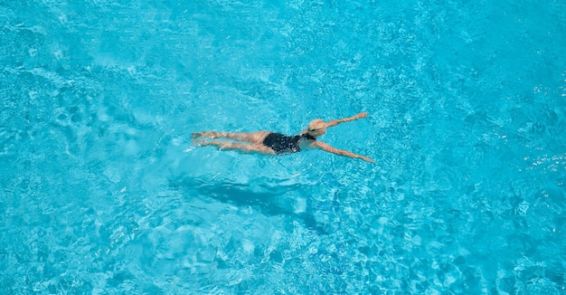 Zenith, ein Blick aus großer Höhe auf eine erwachsene schöne blonde Frau in einem schwarzen Badeanzug, die in einem blauen Pool schwimmt. Freizeit und Urlaub, ein Blick auf den Pool mit einer schwimmenden Europäerin