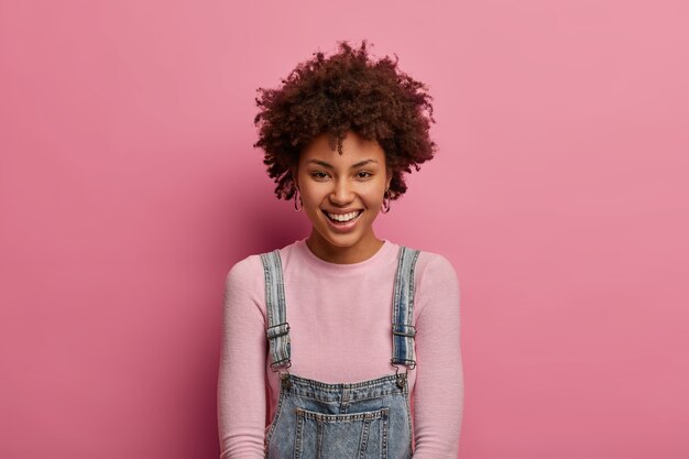 Ziemlich entzückte ethnische Frau mit lockigem Haar, gute Laune, trägt einen Pullover mit Denim-Sarafan, hat einen zarten weiblichen Look, lächelt erfreut, isoliert über der rosa Wand, hat eine saubere Haut