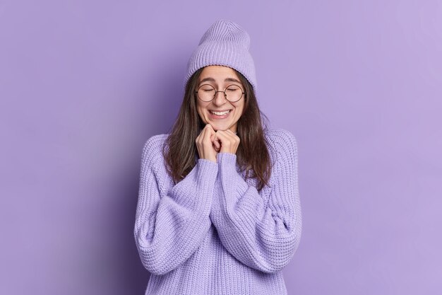 Ziemlich erfreut Brünette Frau hält Hände unter Kinn schließt die Augen und freut sich, etwas Gutes in warmen Winterpullover Hut große Brille gekleidet zu hören.