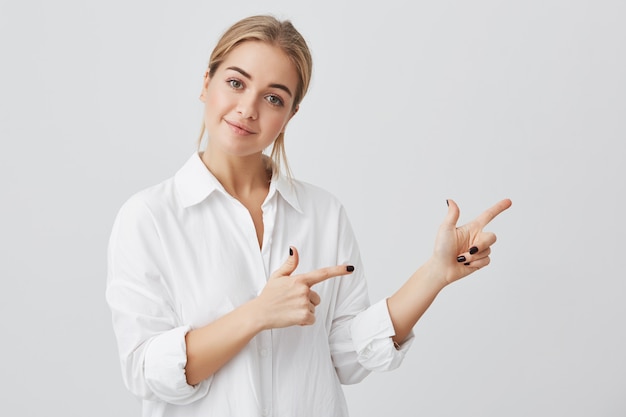 Ziemlich freudig lächelnd weiblich mit hellem Haar, Zeigefinger zeigend, Kopierfläche für Werbeinhalte zeigend. Studioaufnahme des gutaussehenden schönen Mädchens lokalisiert auf Studiowand.