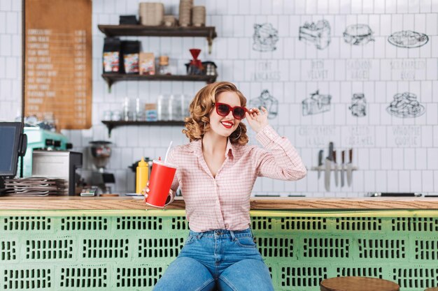 Ziemlich fröhliche Dame mit Sonnenbrille, die mit Sodawasser in der Hand an der Theke sitzt, während sie Zeit im Café verbringt