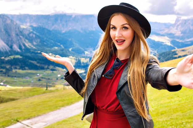 Ziemlich fröhliche junge Touristenfrau, die stilvolle Lederjacke und trendigen Hut trägt und erstaunlichen Blick auf österreichische Alpenberge zeigt