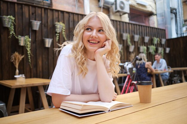Ziemlich fröhliches, lässiges blondes Mädchen, das glücklich mit Buch und Kaffee auf dem Tisch im Innenhof des Cafés wegschaut