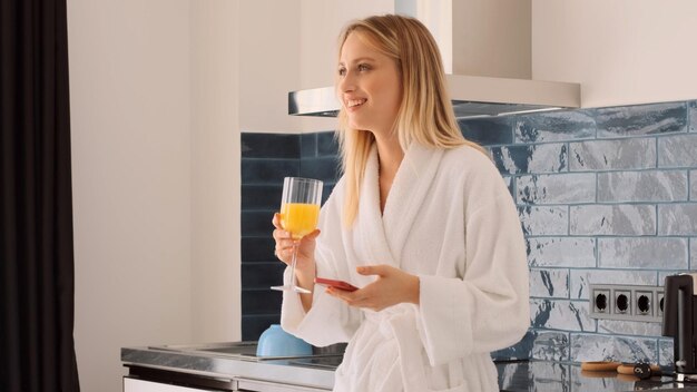 Ziemlich fröhliches Mädchen im weißen Bademantel, das Orangensaft mit Smartphone beim Frühstück in der Küche zu Hause trinkt