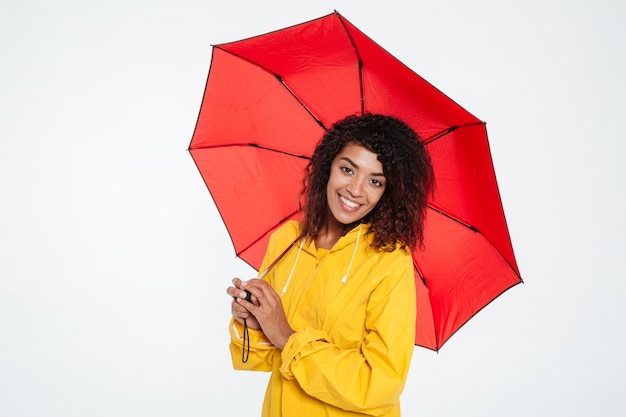 Ziemlich glückliche afrikanische Frau im Regenmantel, der mit Regenschirm aufwirft