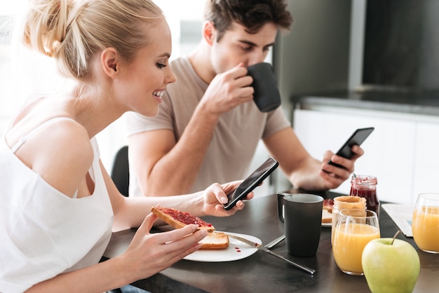 Ziemlich konzentrierte Liebhaber, die ihre Smartphones benutzen, während sie in der Küche frühstücken