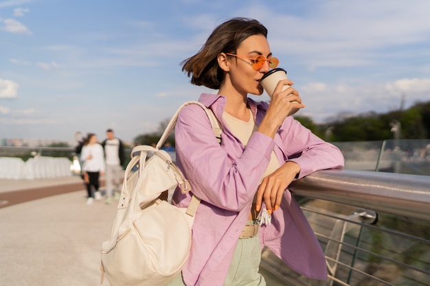 Ziemlich kurzhaarige frau in der stilvollen sonnenbrille, die kaffee genießt, der draußen im sonnigen tag geht