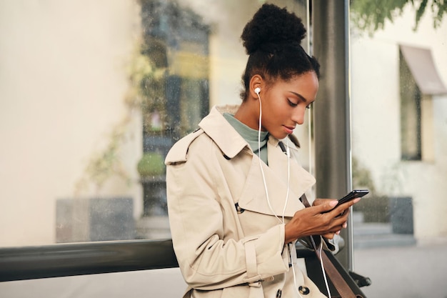 Ziemlich lässiges afroamerikanisches Mädchen in stylischem Trenchcoat und Kopfhörern, das verträumt mit dem Handy an der Bushaltestelle telefoniert