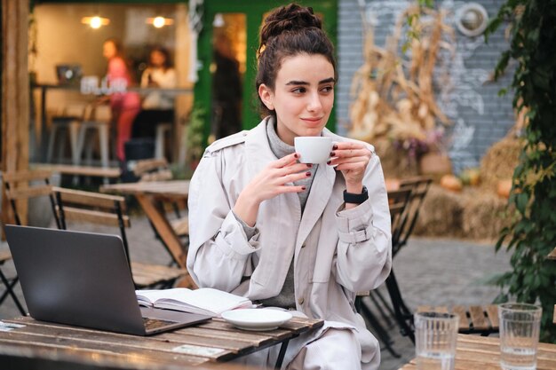 Ziemlich lässiges brünettes Mädchen im Trenchcoat, das Kaffee trinkt und auf dem Laptop im Café auf der Straße der Stadt studiert