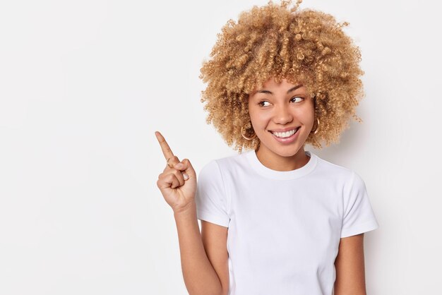 Ziemlich positive Frau mit lockigem Haar lächelt glücklich, zeigt den Zeigefinger in der oberen linken Ecke trägt ein lässiges T-Shirt, das Platz für Ihren Werbeinhalt einzeln auf weißem Hintergrund zeigt.