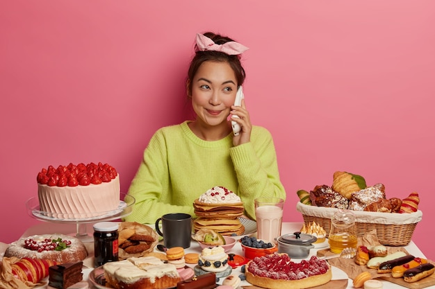 Ziemlich positive koreanische frau hat angenehme telefongespräche, mit süßen süßwaren und kuchen, isst leckeren snack, verwöhnt sich selbst, isoliert über rosa hintergrund