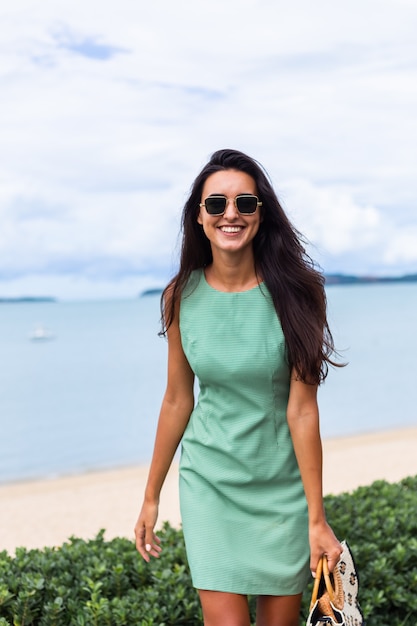 Ziemlich stilvolle glückliche Frau im grünen Sommerkleid mit Tasche, Sonnenbrille im Urlaub tragend, blaues Meer auf Hintergrund