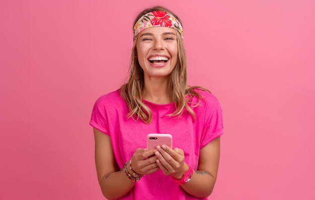 Ziemlich süße lächelnde Frau in rosafarbenem Hemd im Boho-Hippie-Stil mit Accessoires, die emotionalen Spaß lächelt und auf rosafarbenem Hintergrund posiert, isoliert positive Stimmung mit dem Smartphone