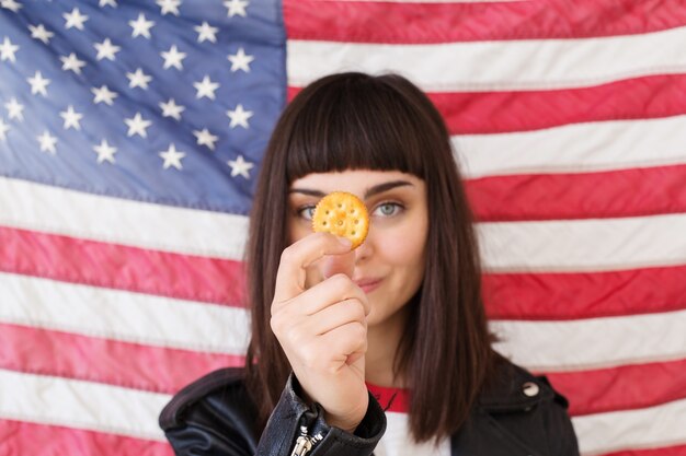 Zierliche weibliche Frau oder Teenager im trendigen Hipster-Outfit isst Posen mit knusprigem Erdnussbutter-Keks-Cracker, typischer traditioneller amerikanischer Snack mit USA-Flagge