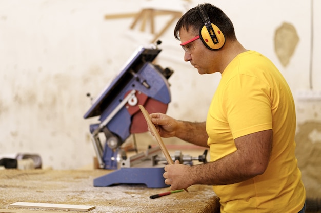 Zimmermann schneidet in seiner Holzwerkstatt ein Stück Holz für Möbel, verwendet eine Kreissäge und trägt eine Schutzbrille und Ohrenschützer.