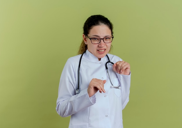 Zimperlicher junger weiblicher Arzt, der medizinisches Gewand und Stethoskop mit den auf olivgrüner Wand isolierten Gläsern trägt