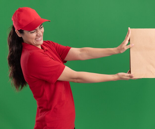 Zimperliches junges Liefermädchen, das Uniform und Kappe trägt, die Papiernahrungsmittelpaket an Kunden lokalisiert auf grüner Wand geben