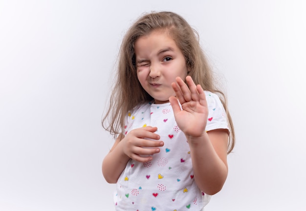 Zimperliches kleines Schulmädchen, das weißes T-Shirt trägt, das Stoppgeste auf lokalem weißem Hintergrund zeigt