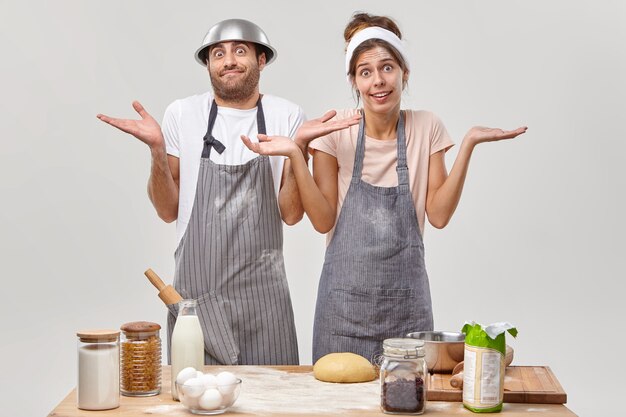 Zögernd befragte junge Frau und Mann zucken mit den Schultern, stehen zusammen in Schürzen, wissen nicht, was sie kochen sollen oder welche Zutaten sie hinzufügen, machen Teig für Kuchen. Bäckereiarbeiter in der Küche bereiten leckeren Kuchen zu