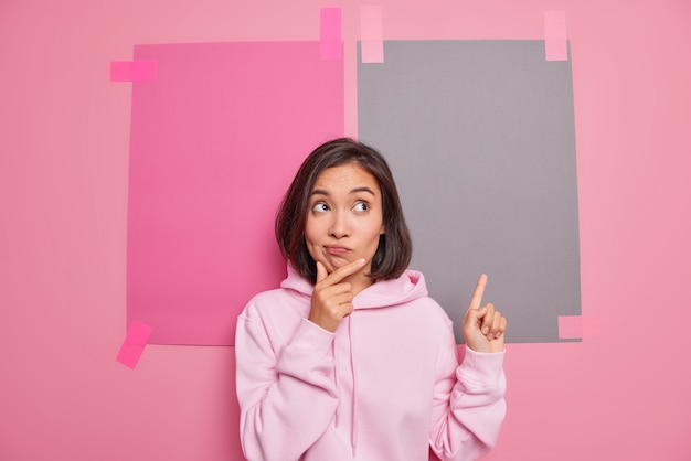 Zögernde brünette junge Asiatin hält Kinntaschen Lippen hat zweifelhaften Gesichtsausdruck zeigt nach oben zeigt Promo vor Top-Verkaufsförderung trägt lässige Hoodie-Posen gegen rosa Wand