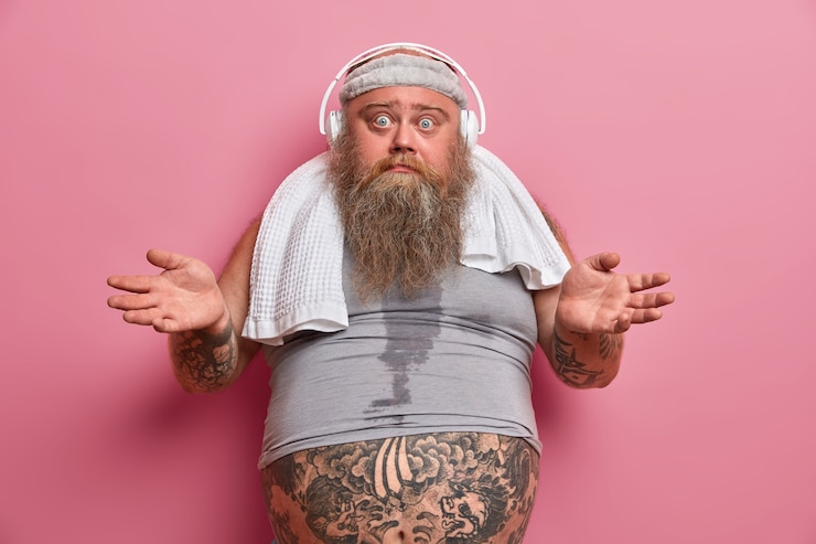 Kostenlose Foto zögernder bärtiger erwachsener mann spreizt die handflächen und sieht verwirrt aus, trainiert regelmäßig zur gewichtsreduktion, hört musik in kopfhörern, trägt ein stirnband und ein untergroßes t-shirt, der tätowierte bauch ragt heraus