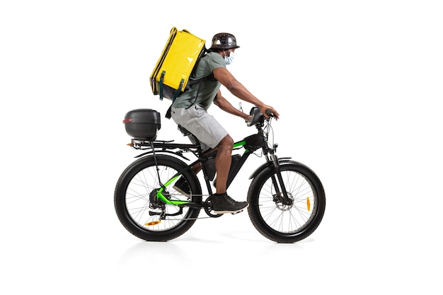 Zu viele Bestellungen. Kontaktloser Lieferservice während der Quarantäne. Mann liefert während der Isolation Essen, trägt Helm und Gesichtsmaske. Nahrungsaufnahme auf dem Fahrrad isoliert auf weißer Wand. Sicherheit. Sich beeilen.