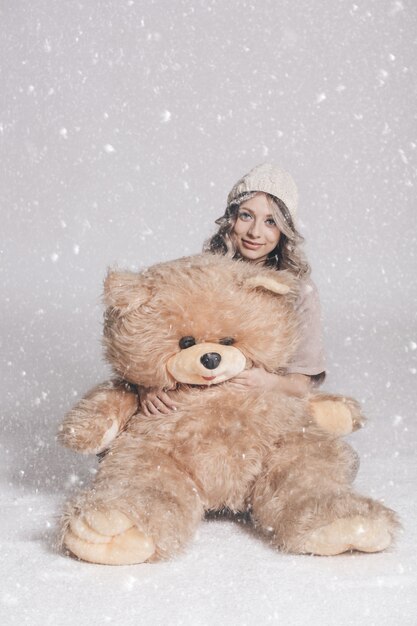 Zufällige lächelnde junge Frau in der gestrickten Kleidung, die großen weichen Teddybären hält, betreffen schneebedeckten Hintergrund