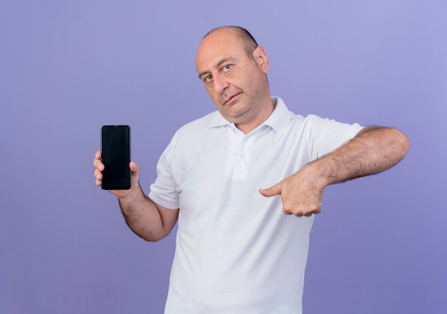 Zufälliger reifer Geschäftsmann, der Kamera betrachtet, die Handy zeigt und auf es zeigt, das Handy lokalisiert auf lila Hintergrund hält