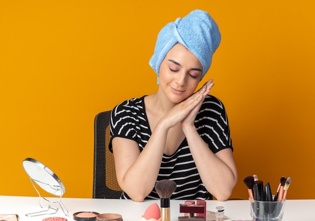 Zufrieden mit geschlossenen augen sitzt das junge schöne mädchen am tisch mit make-up-tools, die haare in ein handtuch gewickelt haben und die schlafgeste einzeln auf der orangefarbenen wand zeigen