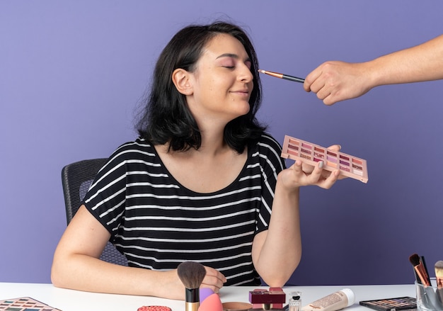 Zufrieden mit geschlossenen Augen sitzt das junge schöne Mädchen am Tisch mit Make-up-Tools, die Lidschatten von jemandem auf der blauen Wand auftragen