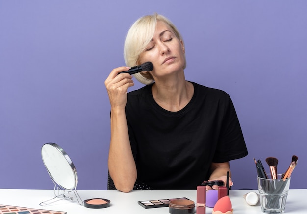 Zufrieden mit geschlossenen Augen sitzt das junge schöne Mädchen am Tisch mit Make-up-Tools, die Puderrouge mit Puderpinsel einzeln auf blauer Wand auftragen