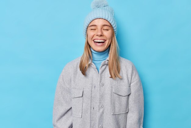 Zufriedene aufrichtige junge europäische Frau hält die Augen geschlossen, lächelt im Großen und Ganzen positive Emotionen aus und trägt eine graue Jacke mit Strickmütze einzeln über blauer Studiowand. Leutekleidung und lustiges Konzept