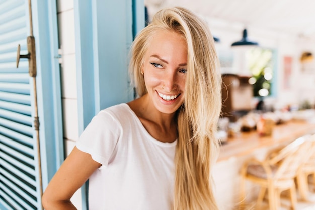 Kostenloses Foto zufriedene blauäugige frau, die spielerisch wegschaut innenfoto eines eleganten gebräunten mädchens mit langen blonden haaren trägt ein weißes t-shirt