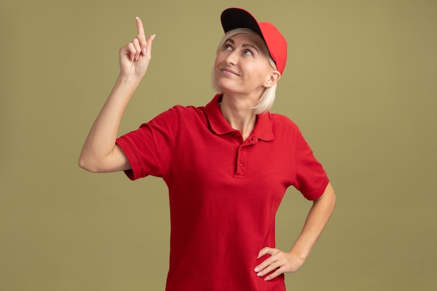 Zufriedene blonde Lieferfrau mittleren Alters in roter Uniform und Mütze, die die Hand auf der Taille hält und nach oben zeigt