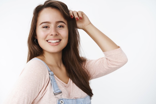 Zufriedene, charmante, freundlich aussehende, attraktive weibliche Brünette, die mit Haaren spielt, lacht und lächelt, als sie auf der linken Seite steht und positive freudige Emotionen über grauer Wand ausdrückt. Platz kopieren