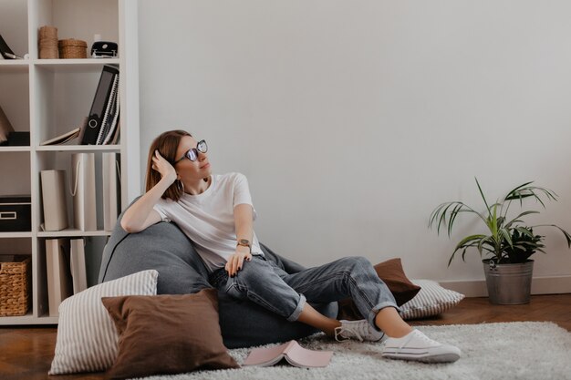 Zufriedene Frau in Jeans und T-Shirt entspannt sitzen im Taschenstuhl gegen Regal mit Ordnern.