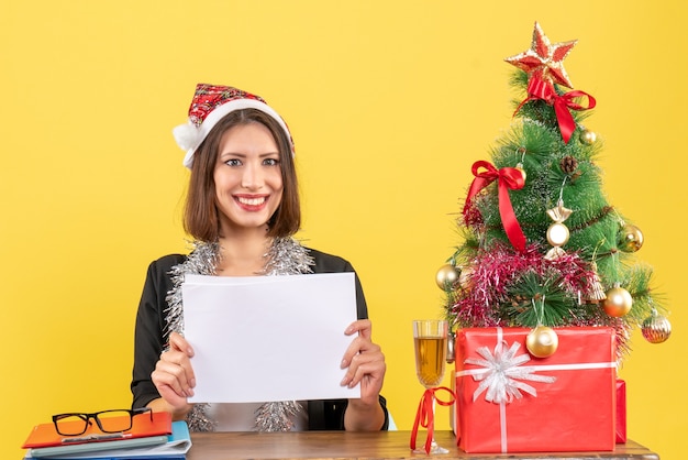 Zufriedene Geschäftsdame im Anzug mit Weihnachtsmannhut und Neujahrsdekorationen, die alleine arbeiten, Dokumente halten und an einem Tisch mit einem Weihnachtsbaum darauf im Büro sitzen