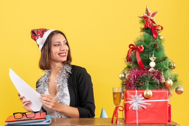 Zufriedene Geschäftsdame im Anzug mit Weihnachtsmannhut und Neujahrsdekorationen, die alleine arbeiten und an einem Tisch mit einem Weihnachtsbaum darauf im Büro sitzen