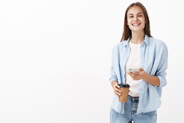 Zufriedene glückliche attraktive kaukasische Frau in der blauen Bluse über T-Shirt halten Pappbecher Kaffee und Handy lächelnd mit Freude glücklich, Pause während der Arbeit im Büro zu haben