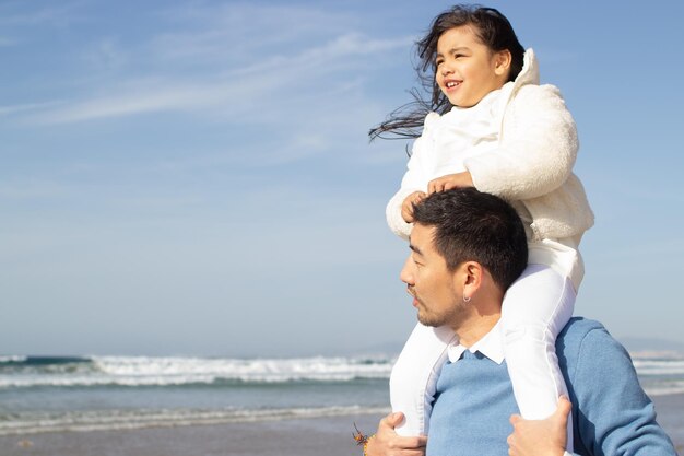 Zufriedene japanische Familie, die am Strand spazieren geht. Tochter reitet auf dem Rücken des Vaters und freut sich. Freizeit, Urlaub, Elternkonzept