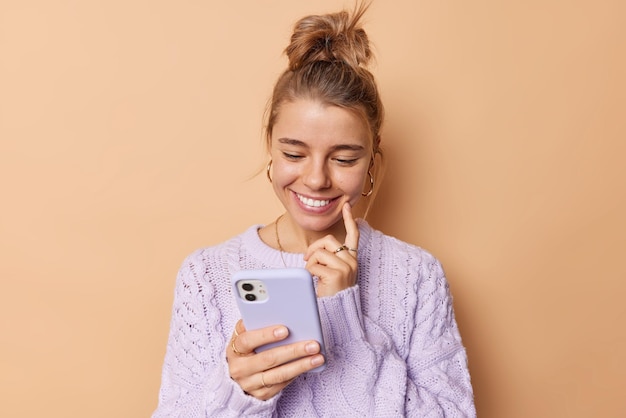 Zufriedene junge frau lächelt glücklich, hält den zeigefinger auf der wange, konzentriert auf das smartphone, liest eine angenehme nachricht und trägt einen lässigen pullover, der über beigem hintergrund isoliert ist. mobilfunktechnologie