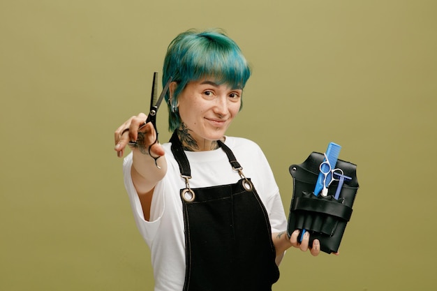 Kostenloses Foto zufriedene junge friseurin in uniform, die eine friseurtasche mit friseurwerkzeugen hält und in die kamera schaut, die eine schere in richtung kamera streckt, isoliert auf olivgrünem hintergrund