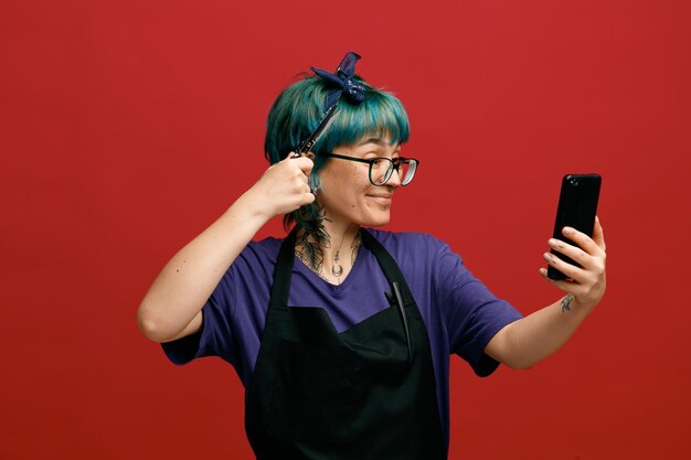 Zufriedene junge Friseurin mit einheitlicher Brille und Stirnband, die das Handy ausstreckt und die Haare mit einer Schere berührt, die ein Selfie mit Haarspange auf ihrer Schürze macht, isoliert auf rotem Hintergrund