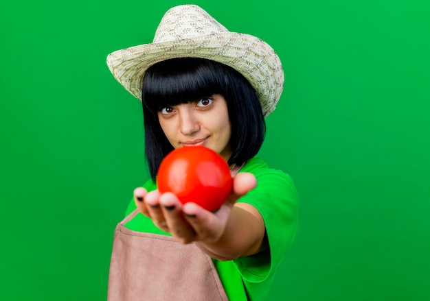 Zufriedene junge Gärtnerin in Uniform mit Gartenhut hält Tomate isoliert aus