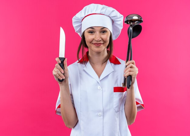 Zufriedene junge Köchin mit Kochuniform, die Messer mit Schöpfkelle auf rosafarbenem Hintergrund hält