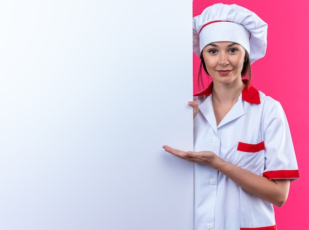 zufriedene junge Köchin mit Kochuniform steht und zeigt mit der Hand auf die nahegelegene weiße Wand, die auf rosafarbener Wand mit Kopienraum isoliert ist