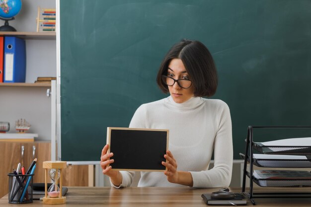 Zufriedene junge Lehrerin mit Brille, die eine Mini-Tafel hält und betrachtet, die am Schreibtisch mit Schulwerkzeugen im Klassenzimmer sitzt