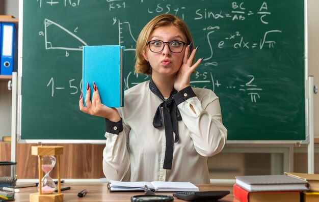 Zufriedene junge Lehrerin mit Brille sitzt am Tisch mit Schulwerkzeugen, die ein Buch im Klassenzimmer halten