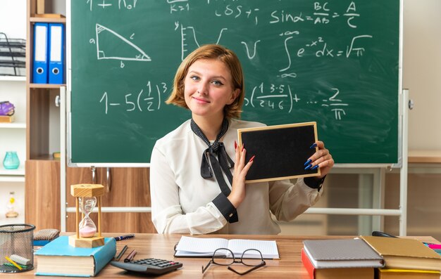 Zufriedene junge Lehrerin sitzt am Tisch mit Schulwerkzeugen, die eine Mini-Tafel im Klassenzimmer halten